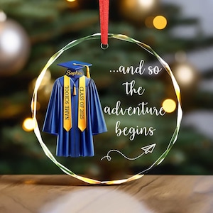 May include: A clear, circular graduation ornament with a blue cap and gown design. The ornament features the text "...and so the adventure begins" and includes a red ribbon for hanging. The gown has "NAME SCHOOL" and "CLASS OF 2025" on the sash.