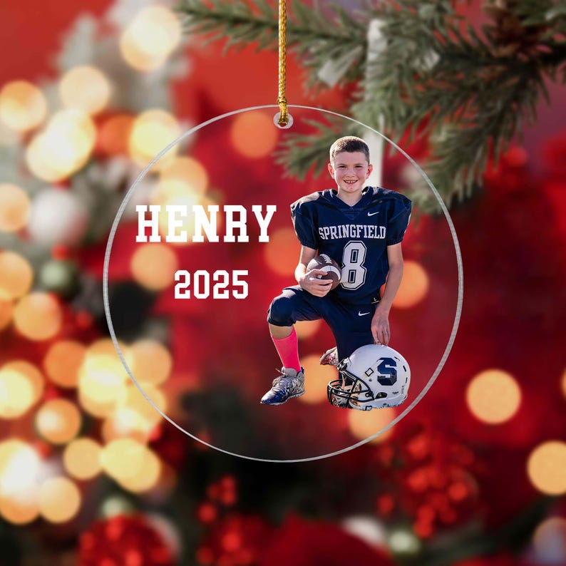 May include: A clear, round ornament with the name "HENRY" and the year "2025" in white text. The ornament features a photo of a young football player in a blue uniform with the number 8, kneeling with a helmet.