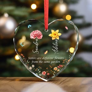 May include: Heart-shaped glass ornament with a red ribbon. The ornament features floral designs, butterflies, and the names "Sandra" and "Adeline." The text reads, "Sisters are different flowers from the same garden."