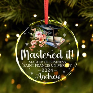 May include: A clear glass Christmas ornament with a white background and a gold rim. The ornament features a watercolor illustration of a graduation cap, diploma, and flowers. The text "Mastered it!" is written in white, followed by "Master of Business Saint Francis University - 2024 - Andrew".