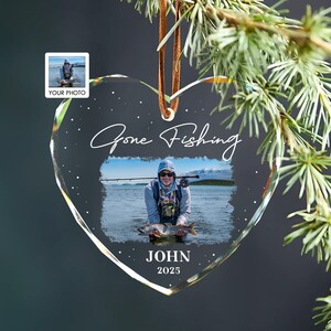 May include: Heart-shaped clear glass ornament with a photo of a person fishing. The ornament has the words "Gone Fishing" in cursive, the name "JOHN", and the year "2025". A small photo is in the upper left corner. A brown leather cord is attached for hanging.