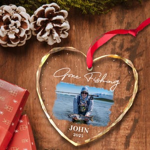 May include: Heart-shaped clear glass ornament with a photo of a person fishing. The ornament has the words "Gone Fishing" and the name "JOHN" with the year "2025". A red ribbon is attached for hanging.