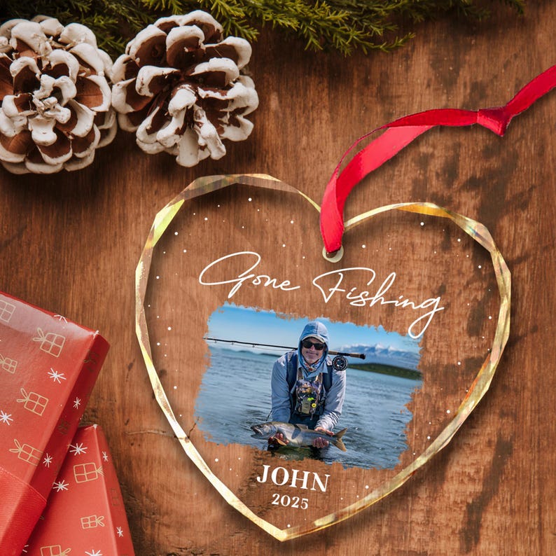 May include: Heart-shaped clear glass ornament with a photo of a person fishing. The ornament has the words "Gone Fishing" and the name "JOHN" with the year "2025". A red ribbon is attached for hanging.
