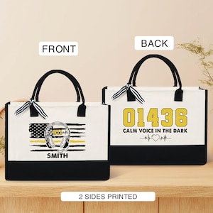 May include: A white canvas tote bag with black handles and a black bottom. The front of the bag features a yellow thin yellow line flag with a 911 headset and the name "SMITH". The back of the bag has the number "01436" in yellow and the text "CALM VOICE IN THE DARK" with a heart beat symbol.