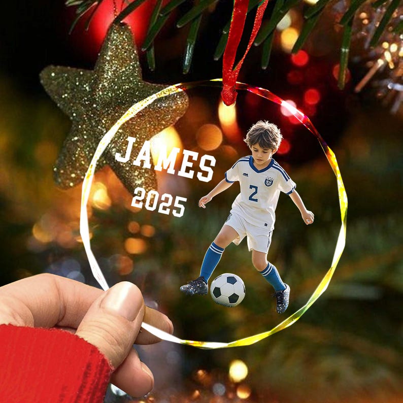 May include: Clear glass ornament with a photo of a young soccer player in a white and blue uniform, kicking a soccer ball. The ornament has the name "JAMES" and the year "2025" printed on it. A red ribbon is attached for hanging.