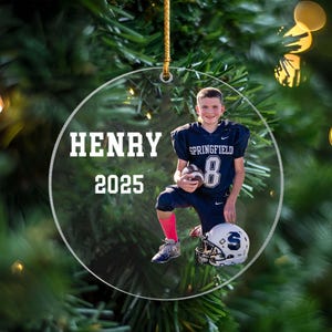 May include: Clear, round Christmas ornament with a photo of a young football player in a navy blue uniform. The ornament has the name "HENRY" and the year "2025" printed on it. The player is kneeling, holding a football and a white helmet.