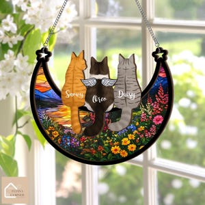 May include: A crescent-shaped cat memorial with three cat figures, each with a name: Snowy, Oreo, and Daisy. The cats are set against a colorful stained-glass background of a landscape and flowers. The piece is suspended by a chain.