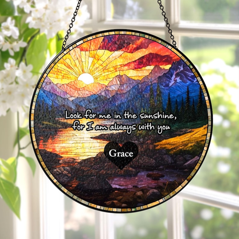 May include: A circular stained glass suncatcher featuring a landscape of mountains, a lake, and a sunset. The text reads, "Look for me in the sunshine, for I am always with you," with a black heart containing the name "Grace" in the center.