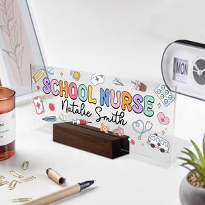 May include: A clear acrylic desk sign with the words "SCHOOL NURSE Natalie Smith" in colorful bubble letters. The sign features medical-themed illustrations and sits in a dark wood base.