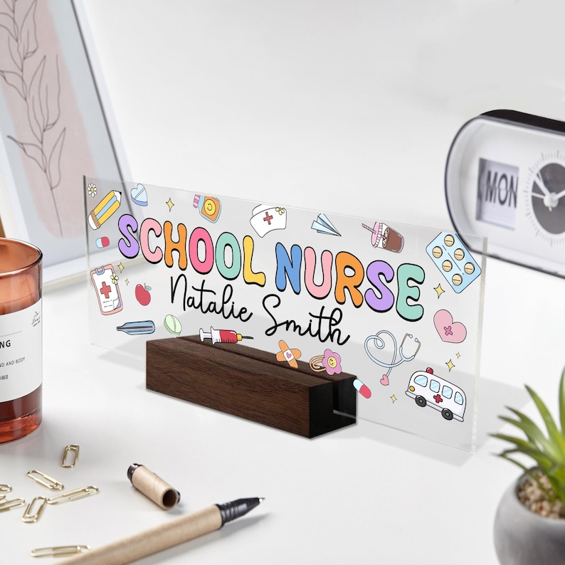 May include: A clear acrylic desk sign with the words "SCHOOL NURSE Natalie Smith" in colorful bubble letters. The sign features medical-themed illustrations and sits in a dark wood base.