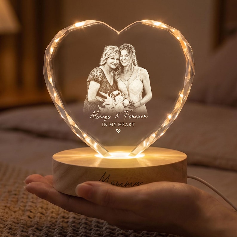 May include: A heart-shaped crystal plaque featuring an engraved monochrome photograph of two individuals, with the text "Always & Forever IN MY HEART" and a small heart symbol. The crystal sits on a round, light-coloured wooden base with warm LED lighting.