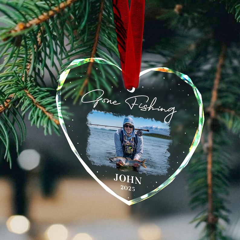 May include: Heart-shaped glass ornament with a photo of a person fishing, the words "Gone Fishing" and the name "JOHN" with the year "2025". The ornament hangs from a red ribbon and is surrounded by green pine needles.