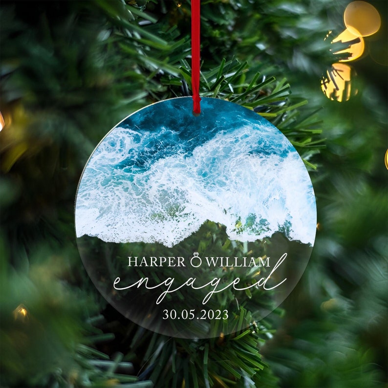 May include: Round clear ornament with a photo of ocean waves crashing on a beach. The text "HARPER & WILLIAM engaged 30.05.2023" is printed on the ornament.