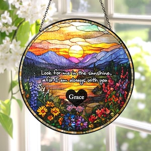May include: A circular stained glass suncatcher featuring a sunset landscape with mountains, a lake, and colorful wildflowers. The text reads, "Look for me in the sunshine, for I am always with you," with a black heart containing the name "Grace."
