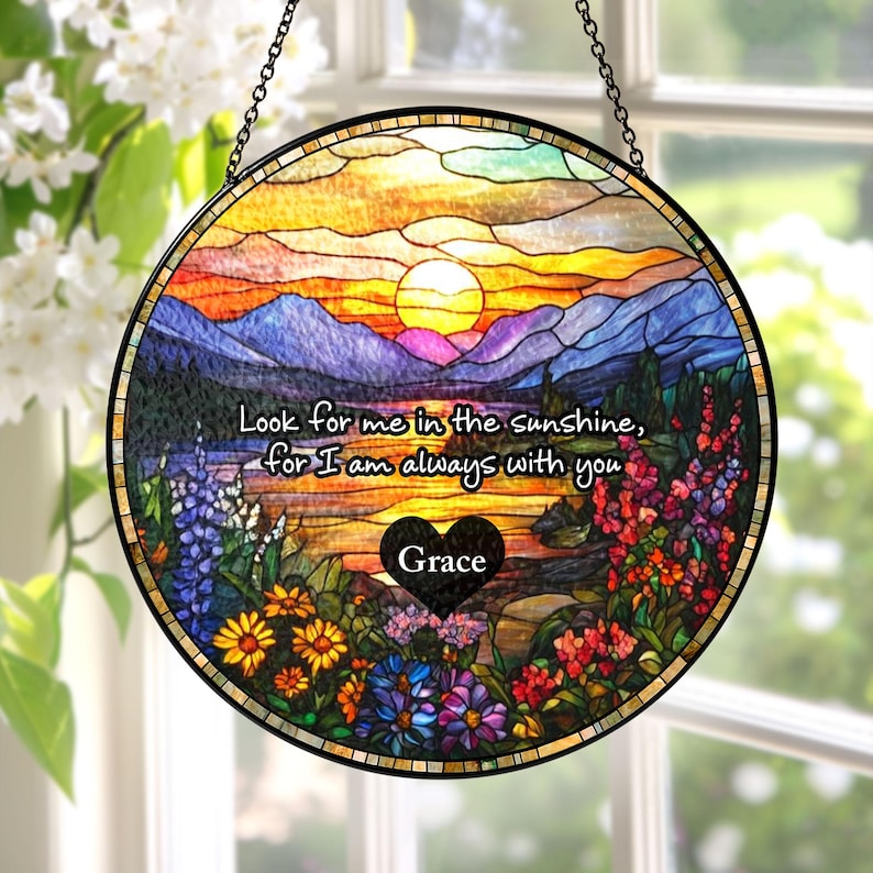 May include: A circular stained glass suncatcher featuring a sunset landscape with mountains, a lake, and colorful wildflowers. The text reads, "Look for me in the sunshine, for I am always with you," with a black heart containing the name "Grace."