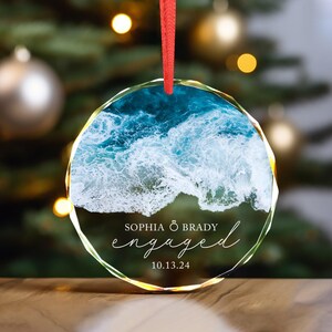 May include: A clear glass ornament with a photo of ocean waves and the text "Sophia & Brady engaged 10.13.24" printed on it.