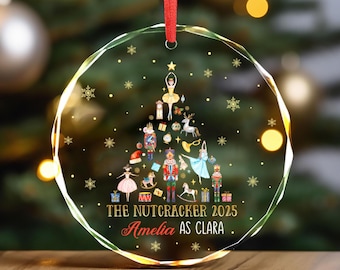 May include: A clear, faceted Christmas ornament with a red ribbon. The ornament features a colorful illustration of a Christmas tree made of Nutcracker characters, snowflakes, and the text "THE NUTCRACKER 2025 Amelia AS CLARA."