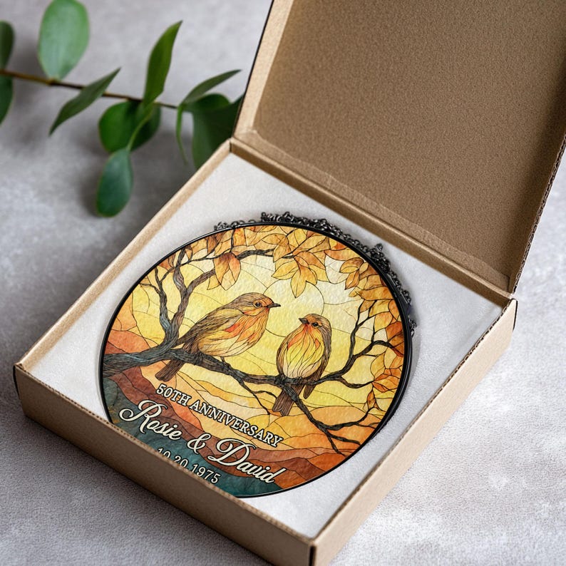 May include: A round stained-glass ornament featuring two birds perched on a branch, set against a warm, autumnal background. The ornament is inscribed with "50TH ANNIVERSARY" and the names "Rosie & David" along with a date. It is presented in a cardboard box.