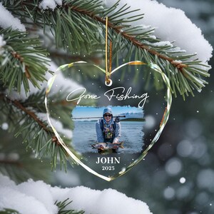 May include: Heart-shaped glass ornament with a gold string, featuring a photo of a person fishing. The ornament has the words "Gone Fishing" and the name "JOHN" with the year "2025" printed on it. The ornament hangs on a snowy evergreen branch.