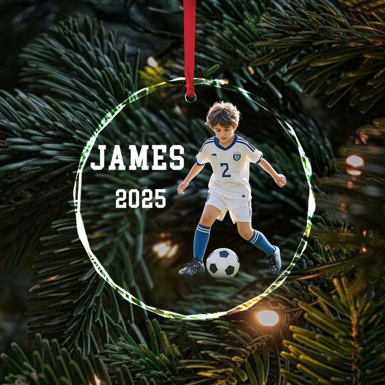 May include: A clear, circular ornament with a photo of a young boy in a soccer uniform kicking a soccer ball. The ornament has the name "JAMES" and the year "2025" printed on it. A red ribbon is attached for hanging.