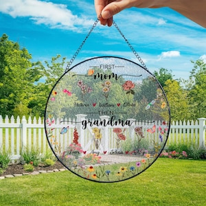 May include: A round, clear glass suncatcher with a black metal frame. It features a floral design with butterflies and the text "First Mom Then Grandma" with names listed below.