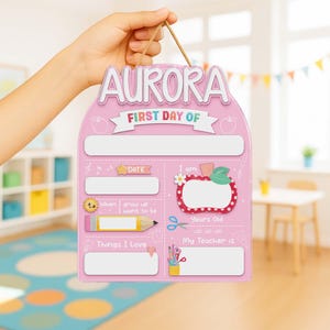 May include: A pink, first day of school sign with the name "AURORA" at the top. The sign has spaces for information like date, age, teacher, and things loved. A brown string is attached for hanging.