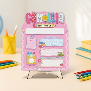 K&ouml;nnte beinhalten: Ein rosa Holzschild mit dem Namen "Camellia" in bunten Buchstaben. Das Schild ist f&uuml;r den ersten Schultag gedacht und enth&auml;lt Felder zum Eintragen von Datum, Alter, Lehrername und W&uuml;nschen. Es wird von einer kleinen Holzstaffelei getragen.