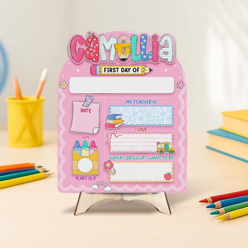 K&ouml;nnte beinhalten: Ein rosa Holzschild mit dem Namen "Camellia" in bunten Buchstaben. Das Schild ist f&uuml;r den ersten Schultag gedacht und enth&auml;lt Felder zum Eintragen von Datum, Alter, Lehrername und W&uuml;nschen. Es wird von einer kleinen Holzstaffelei getragen.