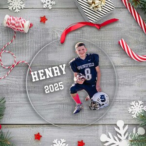 May include: A clear, round Christmas ornament featuring a photo of a person in a football uniform. The ornament has the name "HENRY" and the year "2025" printed on it. Festive decorations like snowflakes, candy canes, and a wrapped gift surround the ornament.