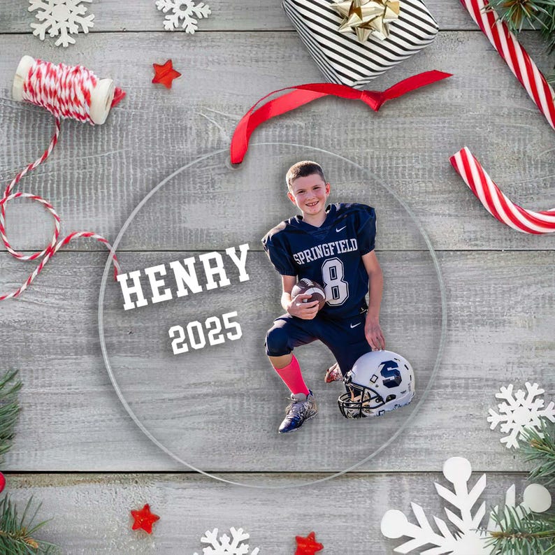 May include: A clear, round Christmas ornament featuring a photo of a person in a football uniform. The ornament has the name "HENRY" and the year "2025" printed on it. Festive decorations like snowflakes, candy canes, and a wrapped gift surround the ornament.