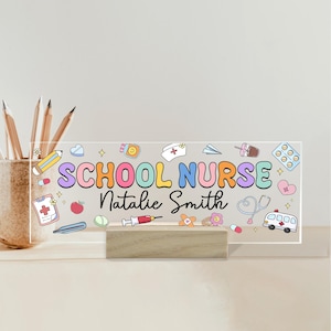 May include: A clear acrylic desk sign with the words "SCHOOL NURSE Natalie Smith" in colorful letters and medical-themed illustrations. The sign is mounted on a wooden base and includes a pencil holder.