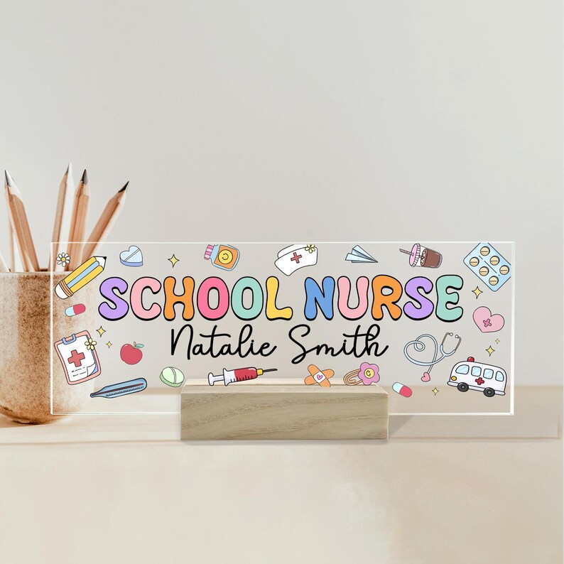 May include: A clear acrylic desk sign with the words "SCHOOL NURSE Natalie Smith" in colorful letters and medical-themed illustrations. The sign is mounted on a wooden base and includes a pencil holder.