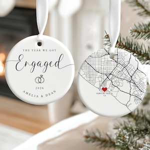 May include: Two round white ceramic ornaments with white ribbon hangers. One features the text THE YEAR WE GOT Engaged, two rings, 2026, and the names AMELIA & DEAN. The other displays a black street map of Newport Beach, CA with a red heart icon.