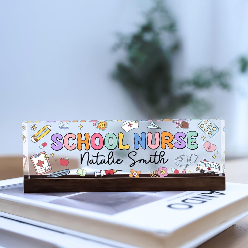 May include: A rectangular acrylic block with the words "SCHOOL NURSE" in colorful bubble letters, along with the name "Natalie Smith." The design includes medical-themed illustrations like a stethoscope, syringe, and pills.
