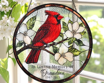 May include: Round stained glass window art featuring a vibrant red cardinal perched on a branch with white flowers. The artwork includes the text "In Loving Memory of Grandma 1947-2025" and is framed with a dark border, hanging by a chain.