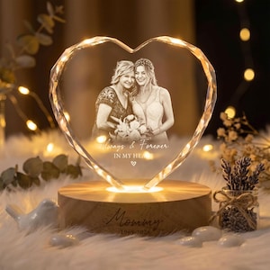 May include: A heart-shaped glass plaque with an etched photograph of two people holding flowers. The text reads Always & Forever IN MY HEART. It sits on a round wooden base engraved with the word Mommy and the years 1969-2026, illuminated by light.