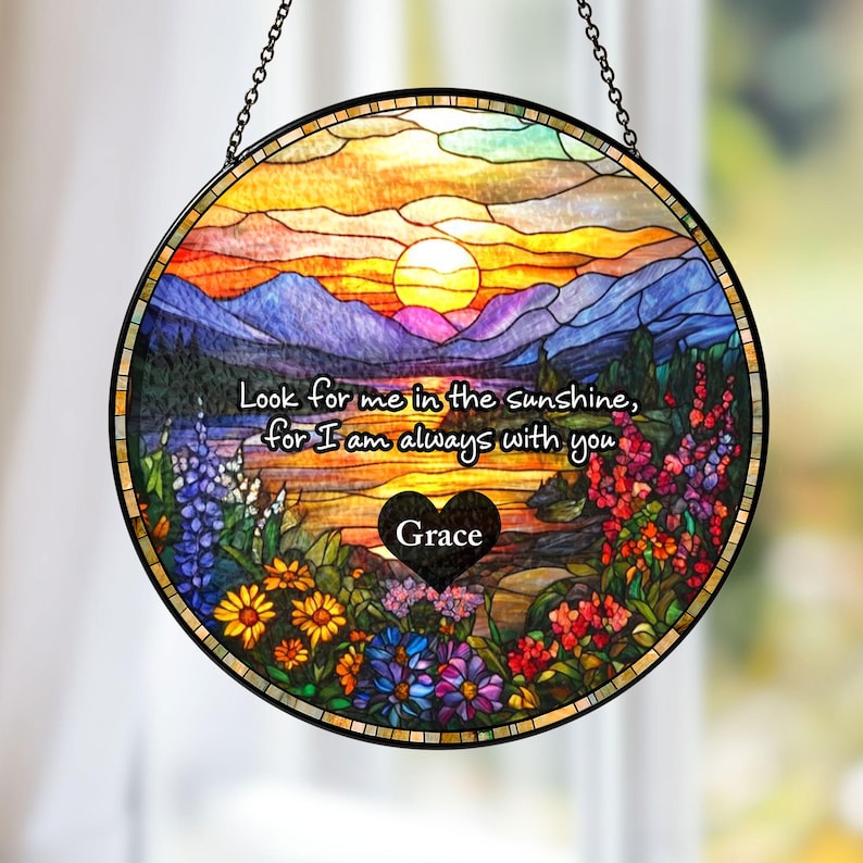 May include: A circular stained glass suncatcher featuring a sunset over mountains and a lake, surrounded by colorful wildflowers. The text reads: Look for me in the sunshine, for I am always with you. A black heart contains the name Grace.