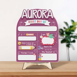 May include: A purple and white first day of school sign with the name "AURORA" at the top. The sign includes spaces for information like date, age, teacher, and things they love. It is supported by a wooden stand.