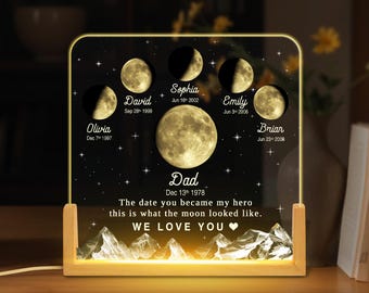 May include: A personalized illuminated moon phase night light with a black background and gold stars. The light features a large moon in the center with smaller moons around it, each with a name and date. The text "Dad Dec 13th 1978" is below the large moon. The text "The date you became my hero this is what the moon looked like. WE LOVE YOU❤" is below the text "Dad".