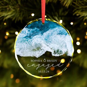 May include: Round glass ornament with a photo of ocean waves and the text "Sophia & Brady engaged 10.13.24"