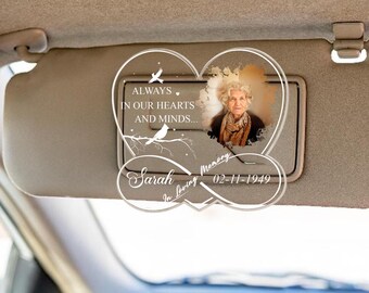 May include: A clear acrylic car visor clip with a heart-shaped infinity symbol. The symbol has the words "In loving memory" and "Always in our hearts and minds..." The clip also has a photo of a woman and the text "Sarah 02-11-1949".