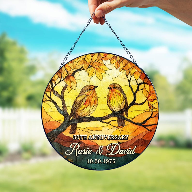 May include: Round stained glass panel featuring two birds perched on a branch, set against a yellow and orange sunset. The panel includes the text "50th Anniversary" and the names "Rosie & David" with the date "10.20.1975". The panel is held by a chain.