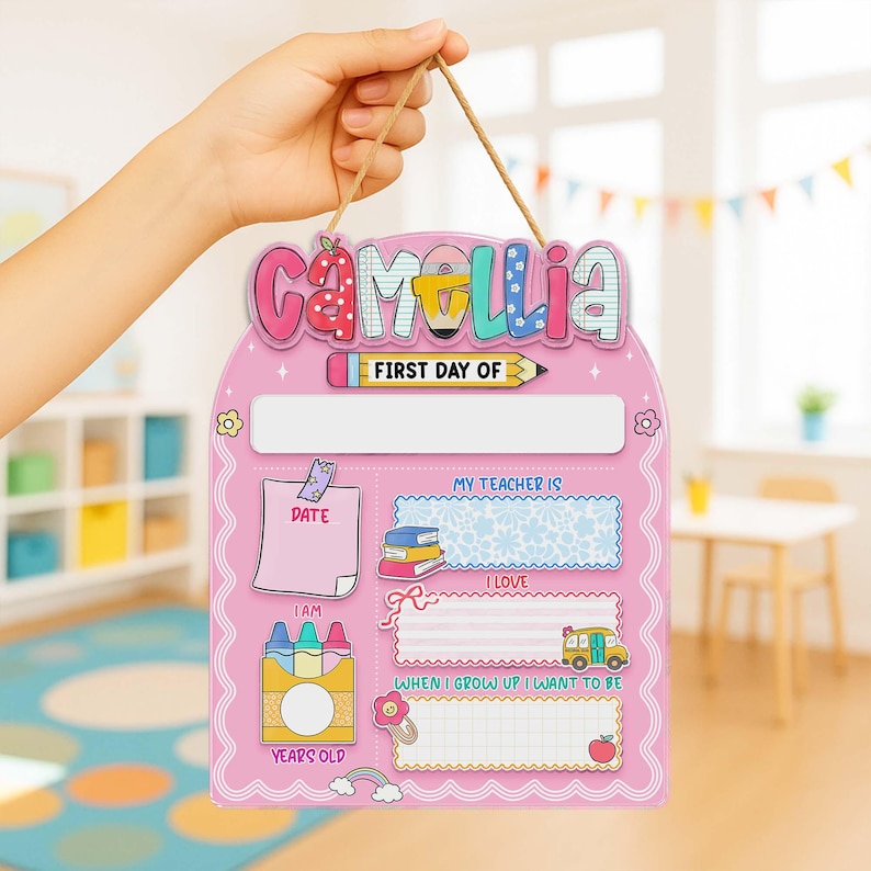 K&ouml;nnte beinhalten: Ein rosa Schulanfangsschild mit dem Namen "Camellia" oben. Das Schild hat Felder f&uuml;r "Datum", "Mein Lehrer ist", "Ich liebe" und "Wenn ich gro&szlig; bin, m&ouml;chte ich sein". Eine Bleistiftgrafik und eine Buntstiftbox sind ebenfalls abgebildet.