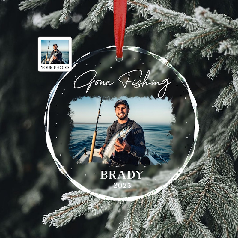 May include: A clear, round Christmas ornament with a photo of a person fishing. The ornament has the words "Gone Fishing", the name "Brady", and the year "2025". A small photo is in the upper left corner. The ornament hangs from a red ribbon.