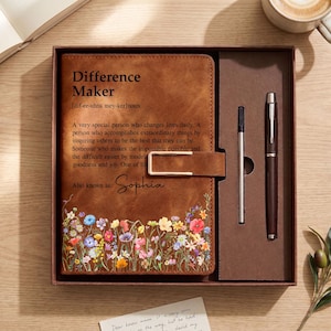May include: A brown leather-effect journal with a gold-tone metal clasp, featuring the printed text "Difference Maker" and a definition, personalised with the name "Sophia" and a colourful floral print at the bottom, presented in a box with a pen.