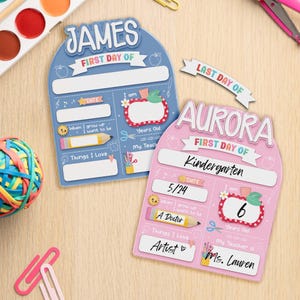 May include: Two colorful first day of school signs. One is blue with the name "JAMES" and the other is pink with the name "AURORA". Both signs have spaces for the date, age, and things they love.