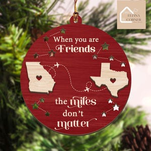 May include: A round, red wooden ornament with the text "When you are Friends the miles don't matter." The ornament features cut-out star shapes and two state outlines with heart details, connected by a dotted line with airplane icons.