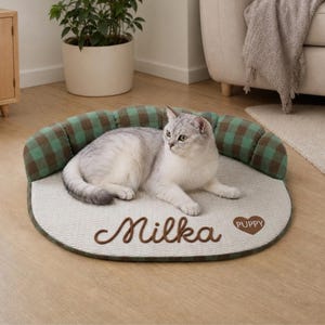 Puede incluir: Un gato gris claro descansando sobre una cama para mascotas con un borde a cuadros verdes y marrones. La cama tiene una base beige texturizada con el nombre Milka bordado y un corazón marrón con la palabra PUPPY en letras blancas.