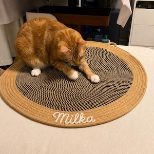 May include: A round, woven rope mat featuring a spiral pattern of tan and black fibres. The mat has a tan outer border and the name Milka written in white cursive script. An orange tabby cat with white paws is sitting on the mat, touching the surface.