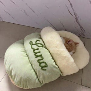 Puede incluir: Un saco de dormir para mascotas de pana verde claro con el nombre Luna bordado en cursiva verde oscuro en la solapa superior. El interior está forrado con piel sintética color crema suave, proporcionando un espacio acogedor para un gato naranja.